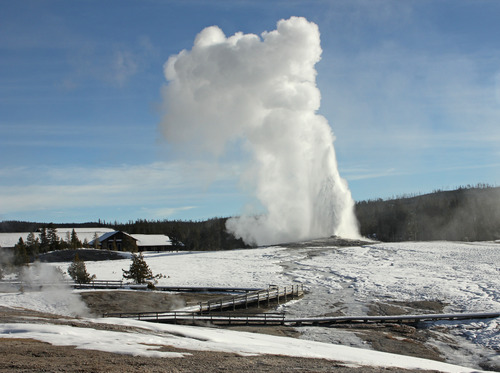 Old Faithful geyser erupting in winter with snow-covered landscape and lodge in background